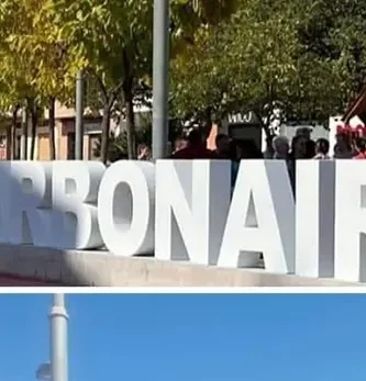 Gran letrero con la palabra 'CARBONAIRE' en una plaza arbolada en Carbonaire, rodeado de personas y vegetación urbana, destacando la participación comunitaria y el entorno cultural del vecindario.