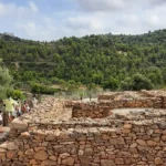 Ruinas arqueológicas en La Vall d'Uixó con un grupo de visitantes observando, rodeados de un paisaje verde y montañoso, destacando el patrimonio histórico y la cultura de la región.