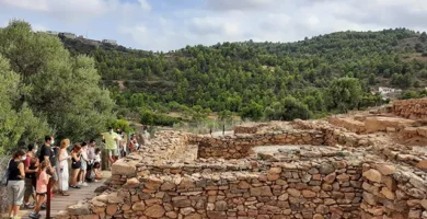 Ruinas arqueológicas en La Vall d'Uixó con un grupo de visitantes observando, rodeados de un paisaje verde y montañoso, destacando el patrimonio histórico y la cultura de la región.