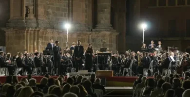Concierto al aire libre de la Ateneo Musical Schola Cantorum La Vall d'Uixó, con la orquesta tocando frente a una iglesia iluminada, y un público numeroso observando el espectáculo nocturno.