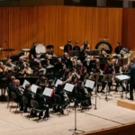 Concierto de la Unión Musical Vallduxense en un auditorio con una orquesta completa, instrumentos de viento y percusión, y un director al frente en Vall d’Uixó.
