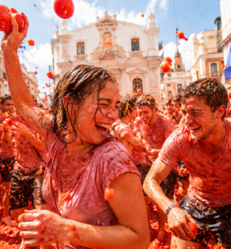 Participantes disfrutando de La Tomatina en Buñol, España, lanzándose tomates en una multitud festiva en medio de una calle llena de edificios con una iglesia al fondo, simbolizando una celebración tradicional española.