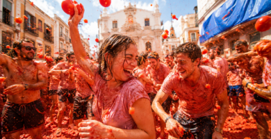 Participantes disfrutando de La Tomatina en Buñol, España, lanzándose tomates en una multitud festiva en medio de una calle llena de edificios con una iglesia al fondo, simbolizando una celebración tradicional española.